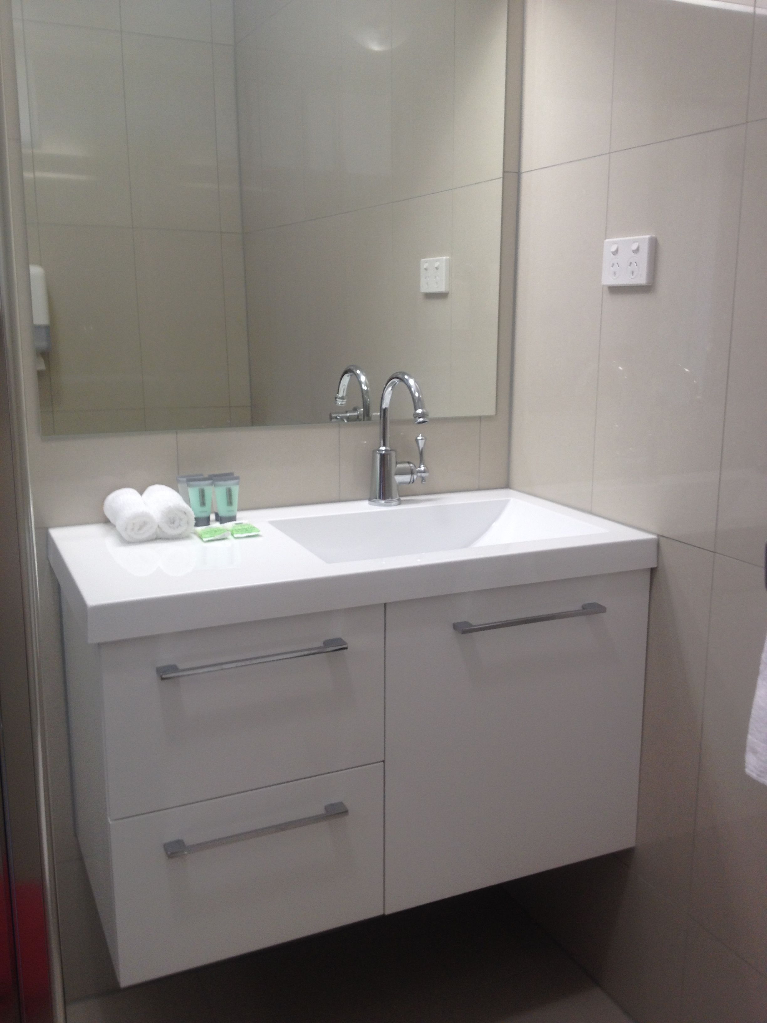 A modern bathroom with a white sink, two faucets, and a mirror above it. There are two drawers and a countertop with toiletries.