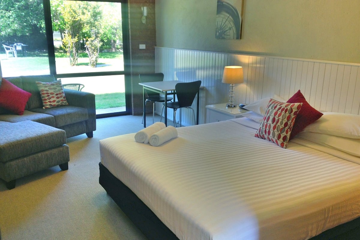 Habitación de hotel con una cama grande, un sofá, un escritorio y una silla. Se ve un ventanal con vistas al exterior.