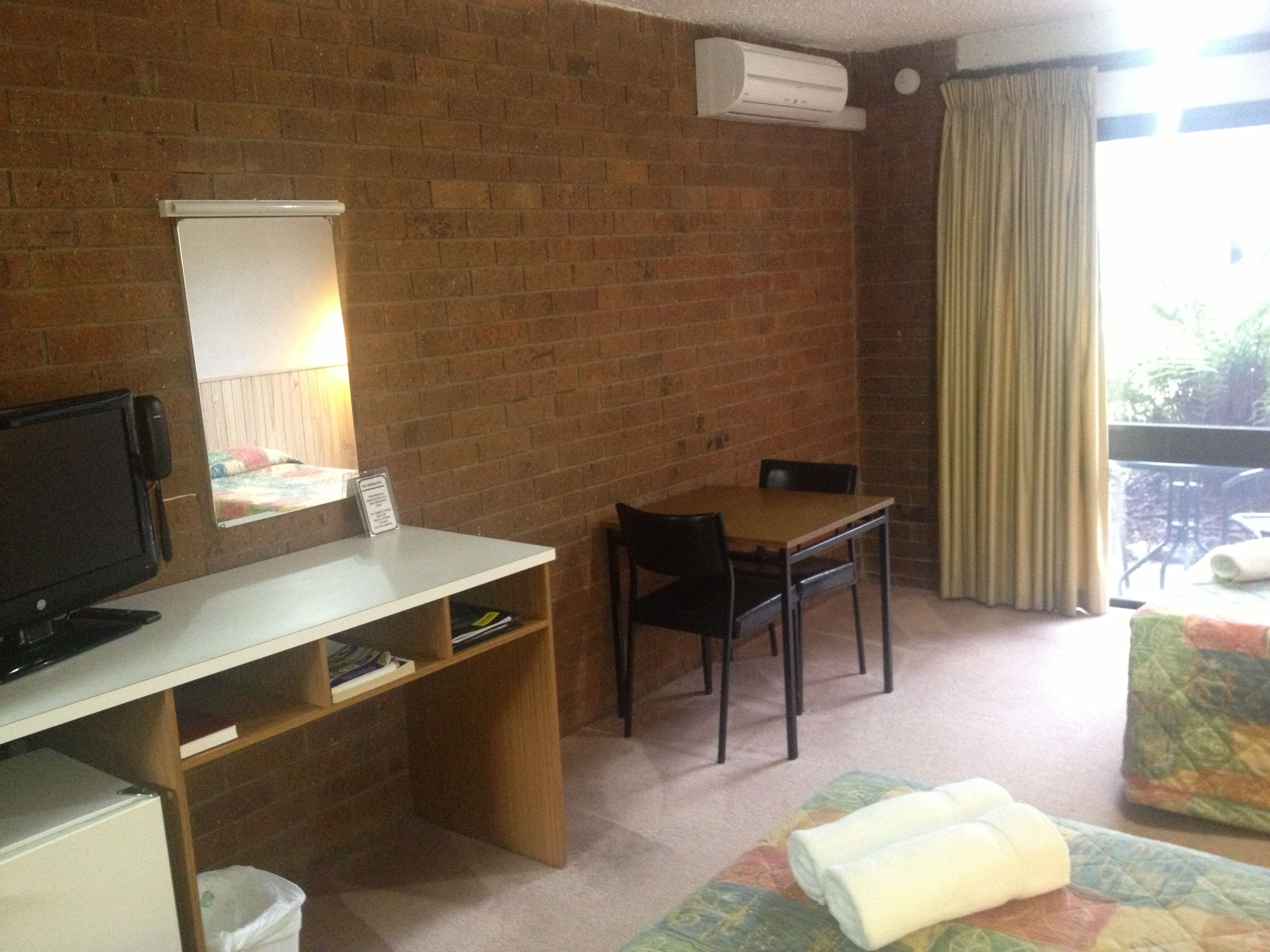 Une pièce avec un mur en briques, un téléviseur posé sur un bureau, une table avec des chaises, une fenêtre avec des rideaux et un lit recouvert d’une courtepointe.
