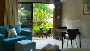 A cozy room with a blue sofa, a small table, and chairs. A large window offers a view of greenery outside. - Motel Sierra (Bright)