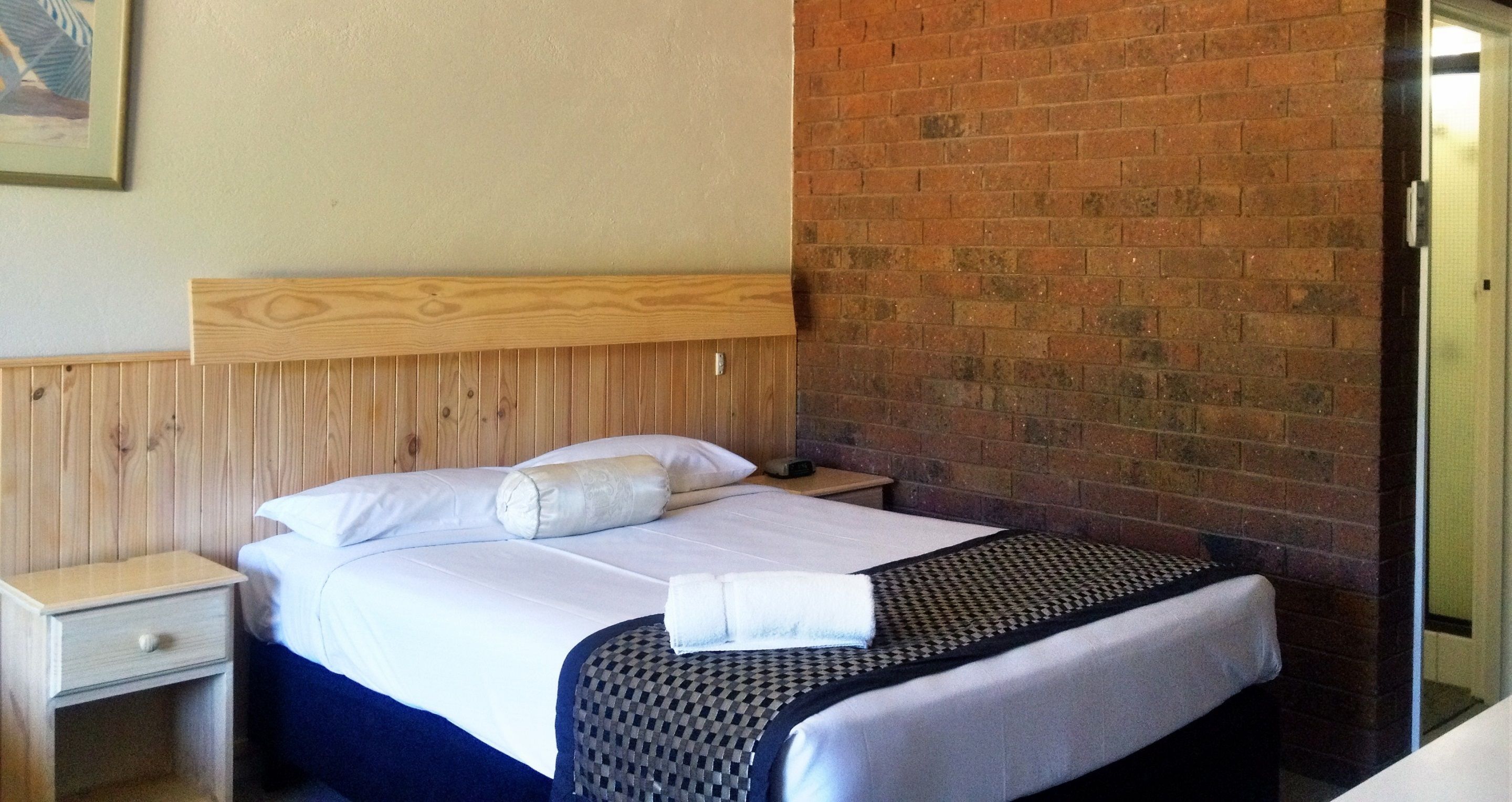 A hotel room with a wooden headboard, a brick wall, a bed with white linens, a nightstand, and a patterned throw blanket.