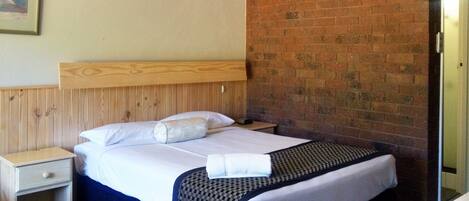 A hotel room with a wooden headboard, a brick wall, a bed with white linens, a nightstand, and a patterned throw blanket.