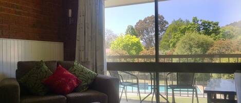 A living room with a sofa, ottoman, and a view of a pool and outdoor seating area through large windows.