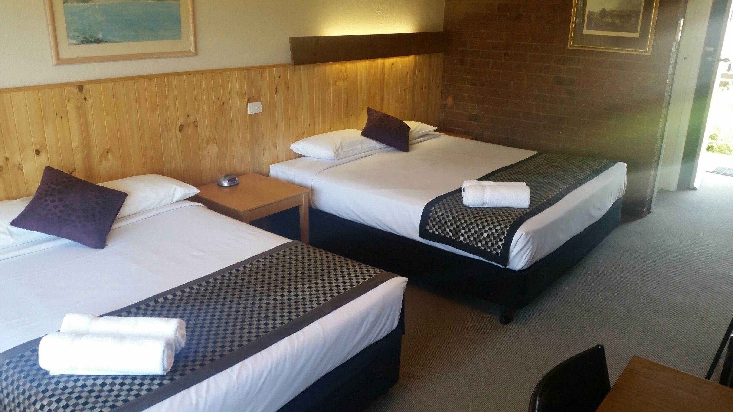 A hotel room with two single beds, each with a pillow and a rolled towel. There is a wooden headboard, a small table with a lamp, and a painting on the wall.