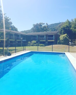 A swimming pool with a clear blue water surface, surrounded by a black metal fence. In the background, there is a multi-story building with balconies and air conditioning units.