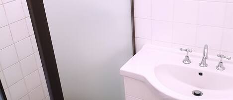 Un baño con un lavabo blanco, dos grifos, un espejo sobre el lavabo y una zona de ducha con puerta de cristal.