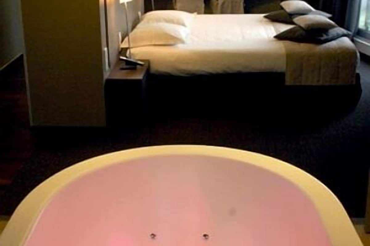 Une chambre d’hôtel dotée d’une grande baignoire moderne, d’un lit parfaitement fait et d’une table de chevet avec une lampe.