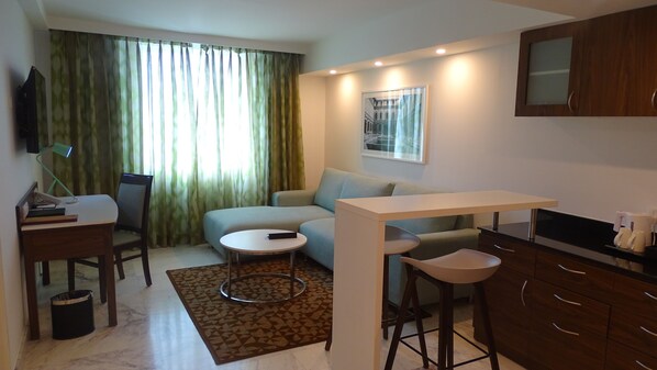 A compact living space with a sofa, a small round table, a TV, a desk with a lamp, and a kitchen area with a coffee machine. - Fairfield by Marriott Pune Kharadi (Pune)
