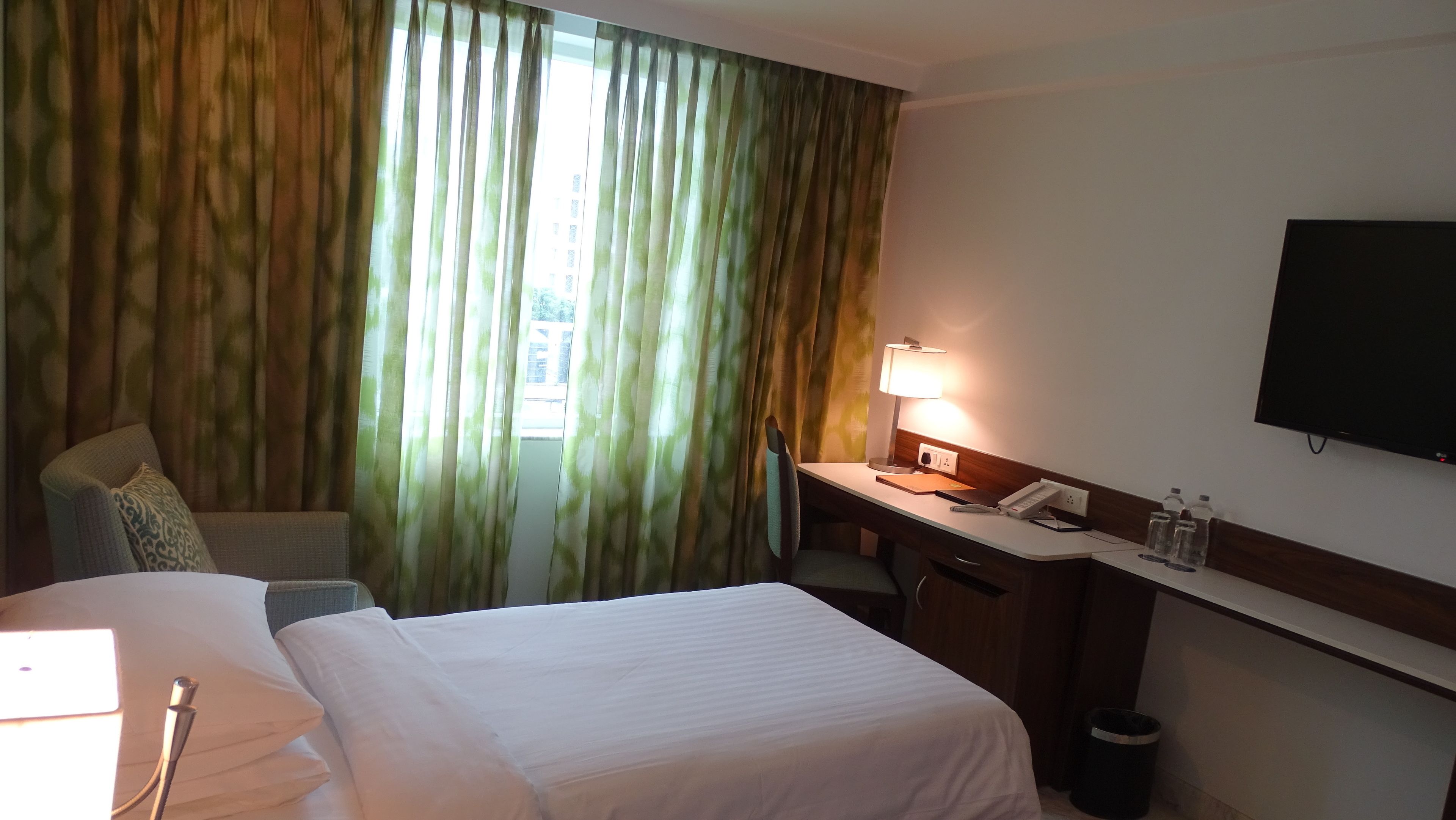 Una habitación de hotel con cama, escritorio, silla, lámpara, televisión y cortinas.