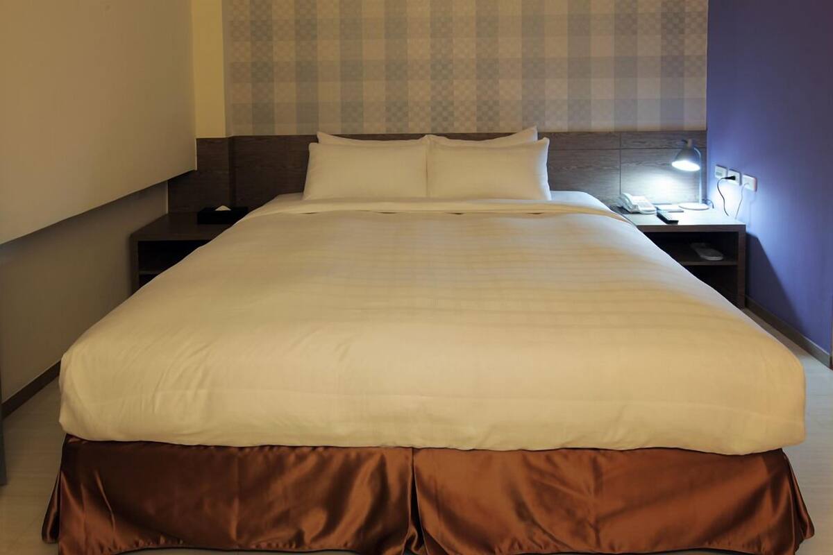 A neatly made bed with white and brown bedding, a nightstand with a lamp, and a patterned wall in the background.
