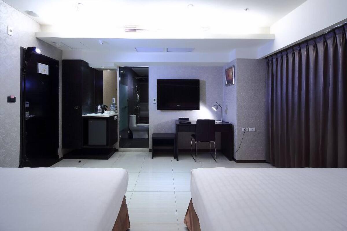 A hotel room with two beds, a desk, a chair, a TV, and a bathroom visible through an open door.