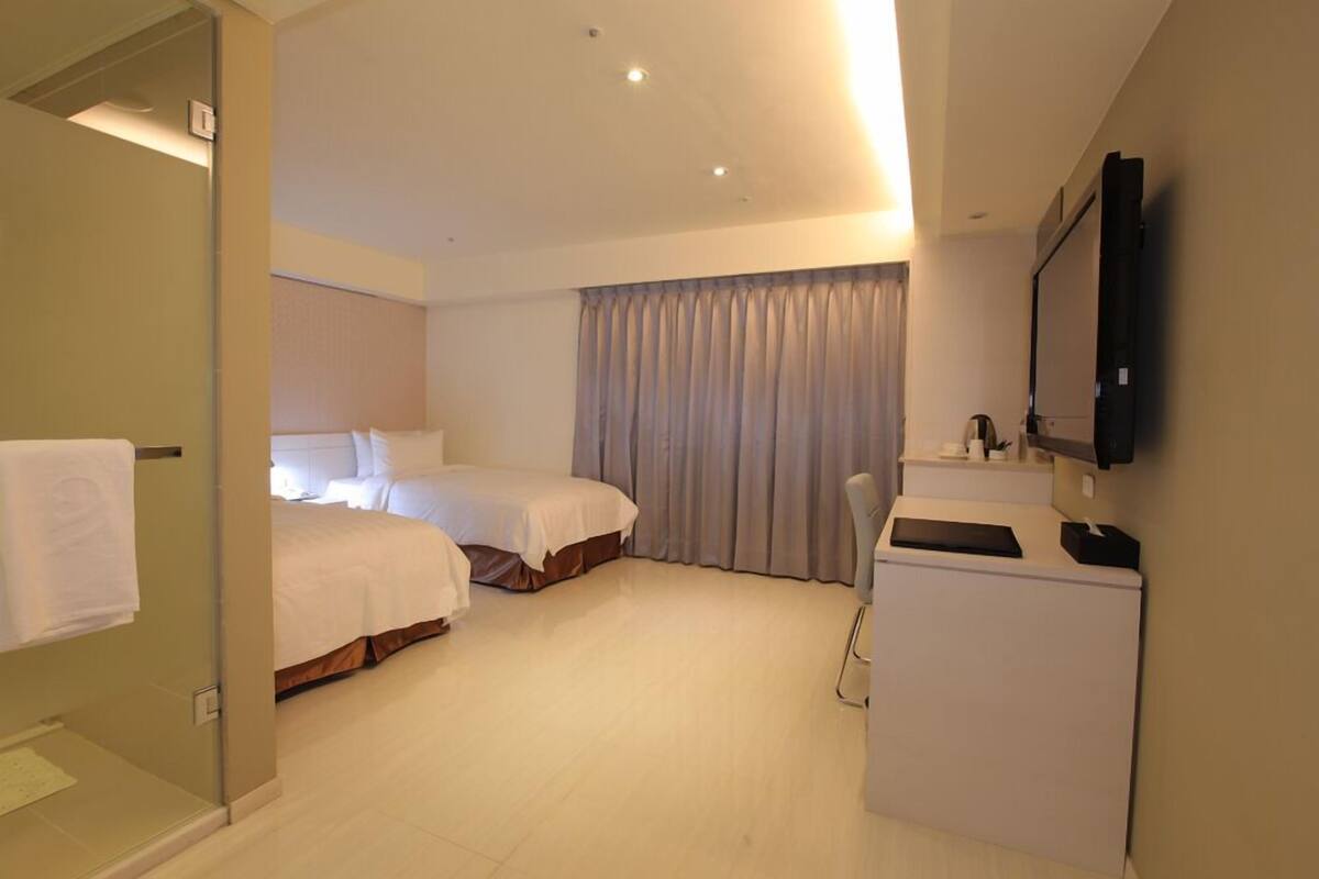 A hotel room with two beds, a desk, a TV, and a shower area.
