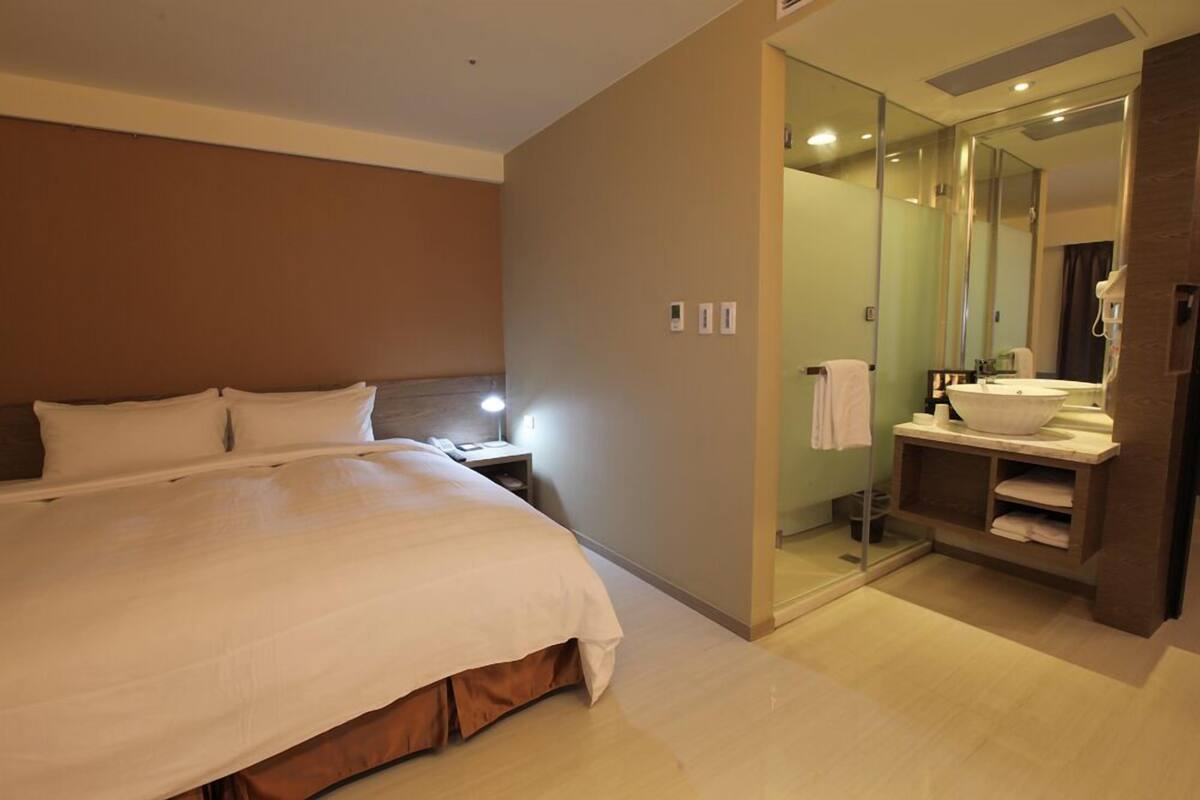 A modern hotel room with a large bed, a bedside table with a lamp, a bathroom with a sink and mirror, and a walk-in closet.