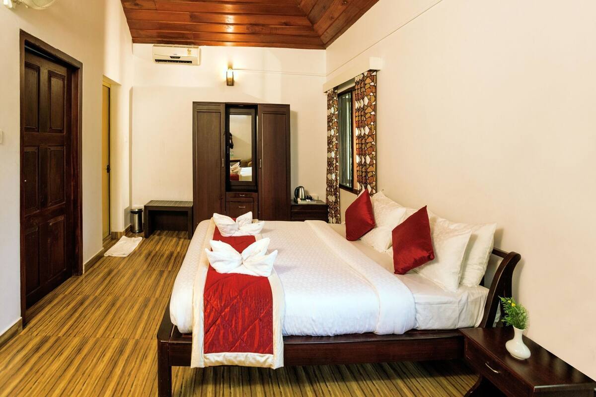 A hotel room with a wooden ceiling, a bed with white bedding and red pillows, a bedside table with a plant, and a mirror on the wall.