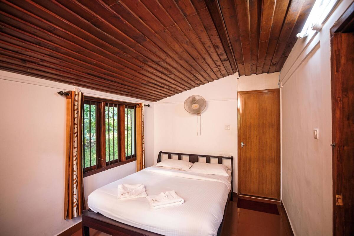 A bedroom with a wooden ceiling, a bed with white linens, a window with curtains, a wall fan, and a wooden door.
