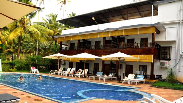 A swimming pool with lounge chairs and umbrellas, surrounded by a building with balconies and a garden. - Raja Park Beach Resort (Varkala)