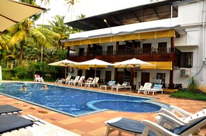 A swimming pool with lounge chairs and umbrellas, surrounded by a building with balconies and a garden. - Raja Park Beach Resort (Varkala)