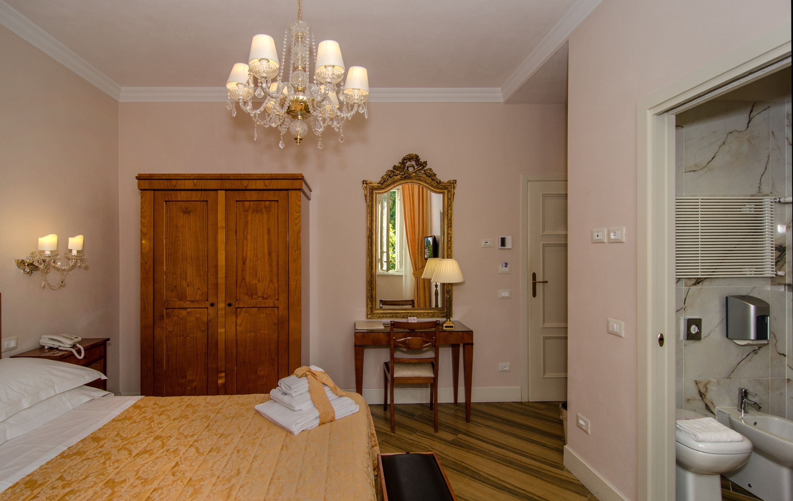 Quarto de hotel com cama, guarda-roupa de madeira, secretária com cadeira, espelho, candelabro e banheiro com pia e vaso sanitário.
