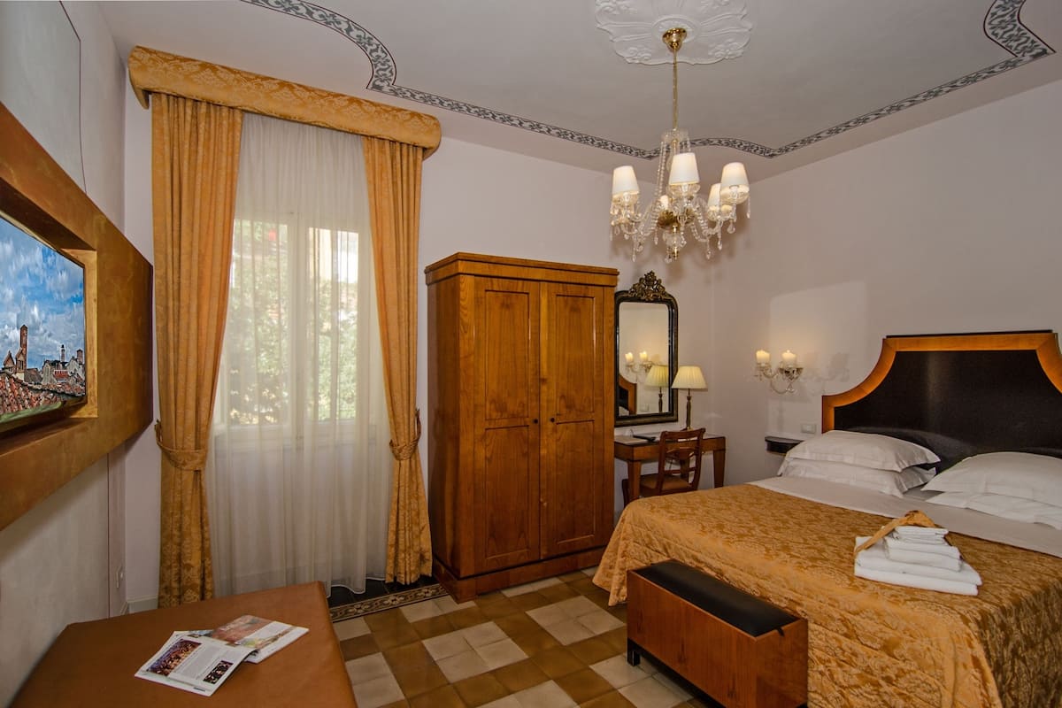 A hotel room with a bed, a wooden wardrobe, a desk with a chair, a chandelier, and a painting on the wall.