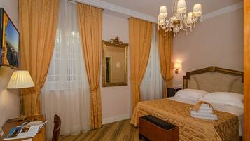 A hotel room with a bed, a desk with a lamp, a chair, a bedside table with a lamp, and a chandelier.