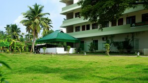 A hotel with a green roof, multiple floors, and a surrounding grassy area with palm trees. - Cosy Regency (Ambalapuzha)