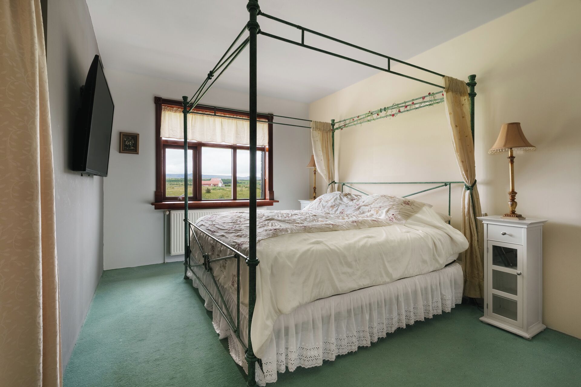 A four-poster bed with a view of the countryside, a bedside table with a lamp, and a radiator.