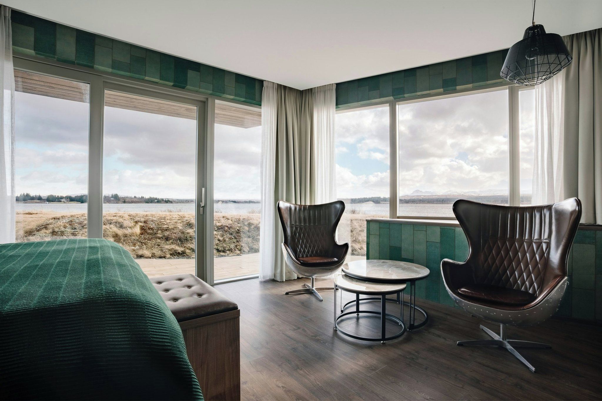 A room with a bed, two leather chairs, a small round table, and a view of a natural landscape through large windows.