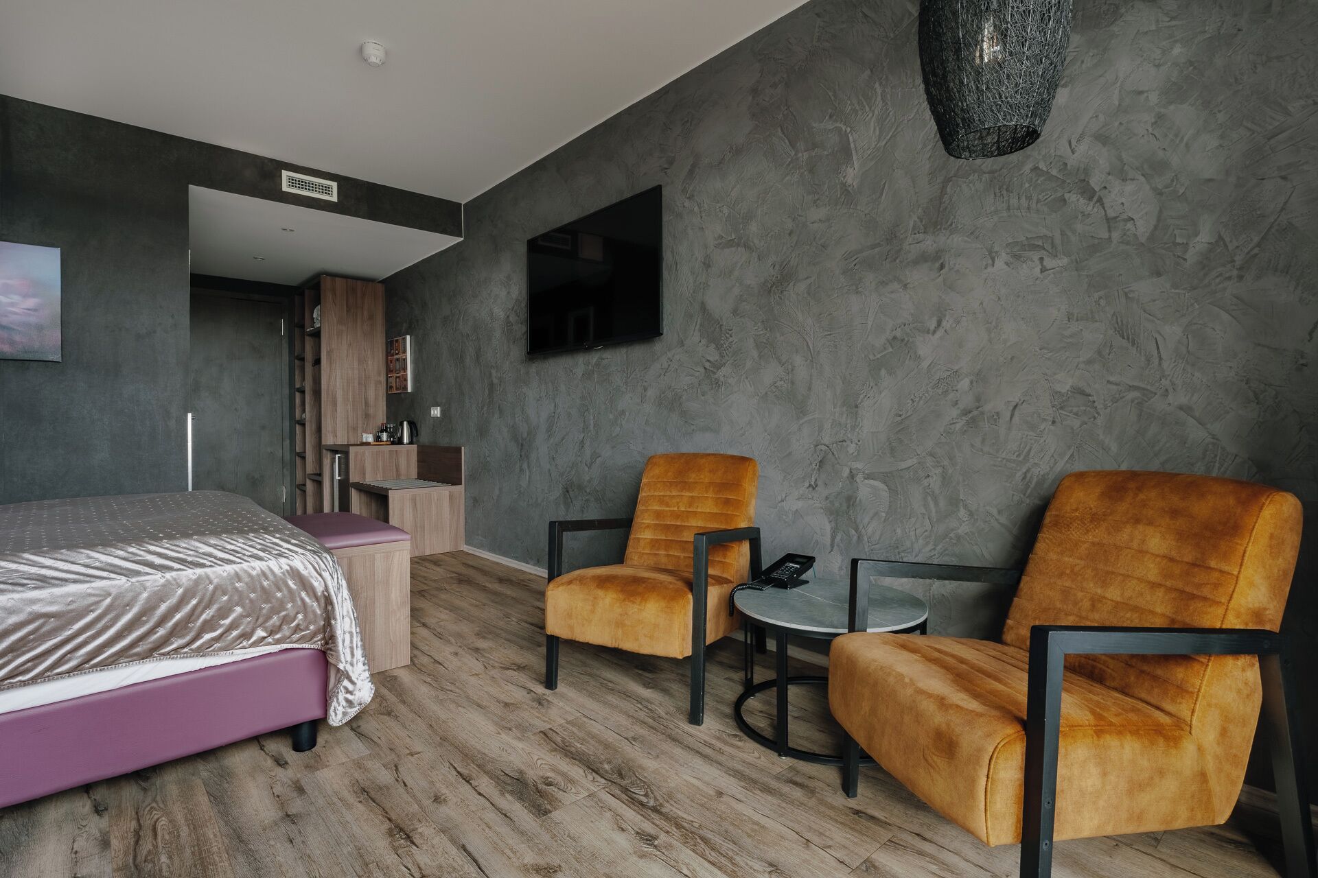 Ett modernt hotellrum med en säng, två fåtöljer, ett litet bord, en TV och ett litet kök.