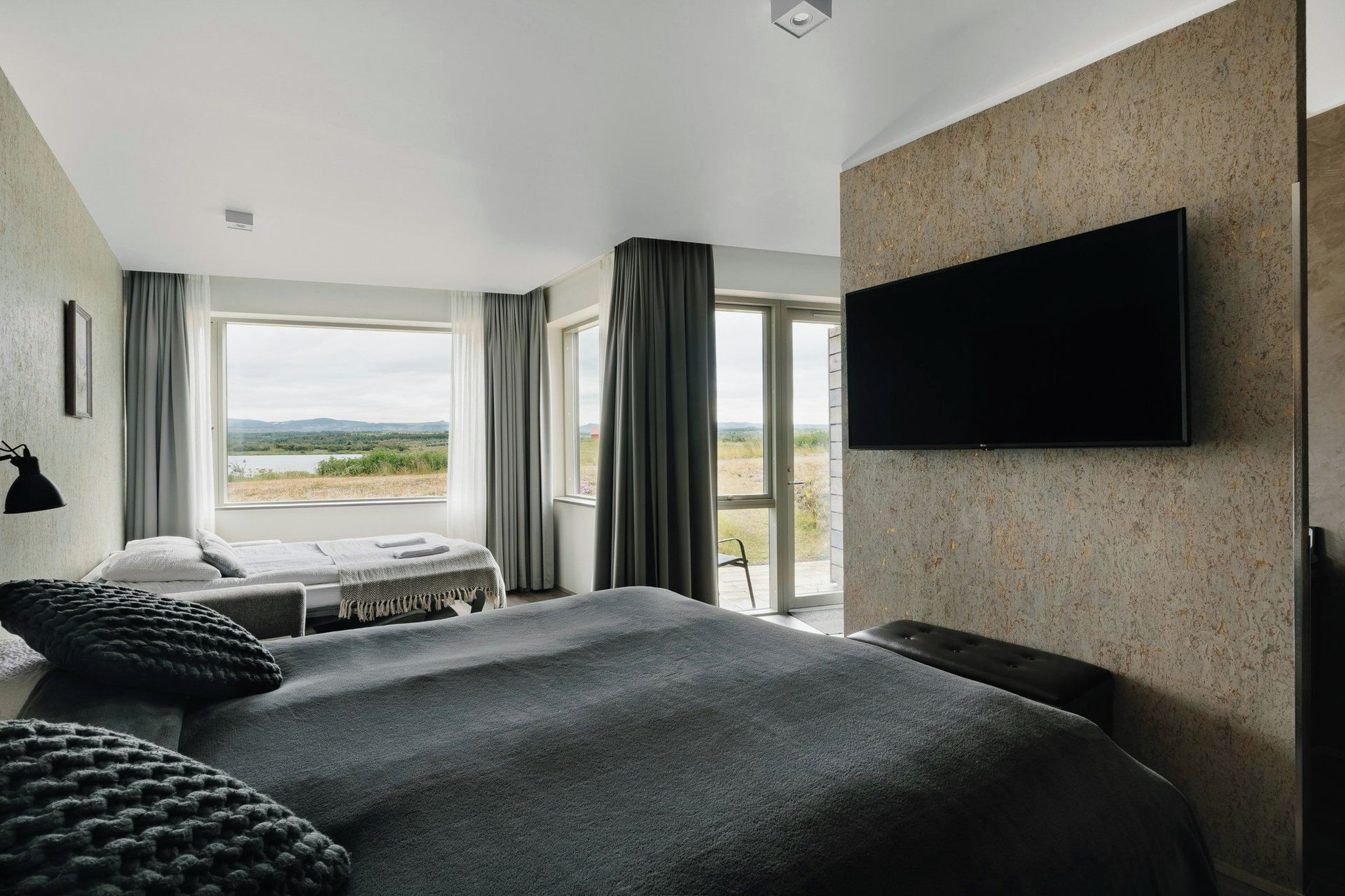 A modern hotel room with a large bed, a flat-screen TV mounted on the wall, and a view of the outdoors.