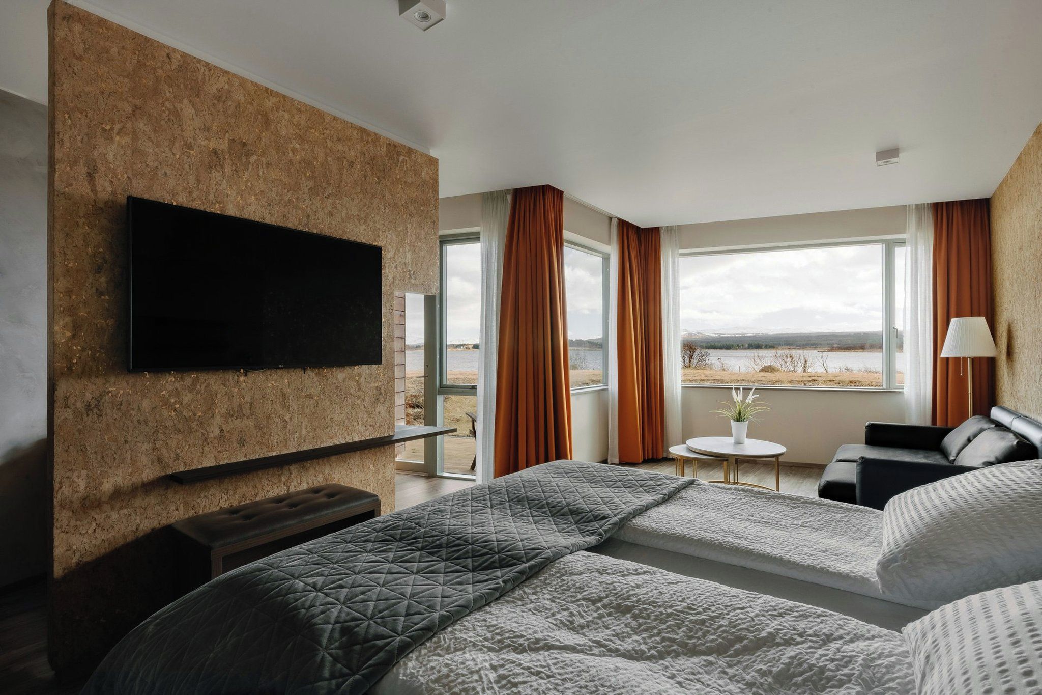 A hotel room with a large bed, a flat-screen TV mounted on a textured wall, a seating area with a table, and a view of a lake.