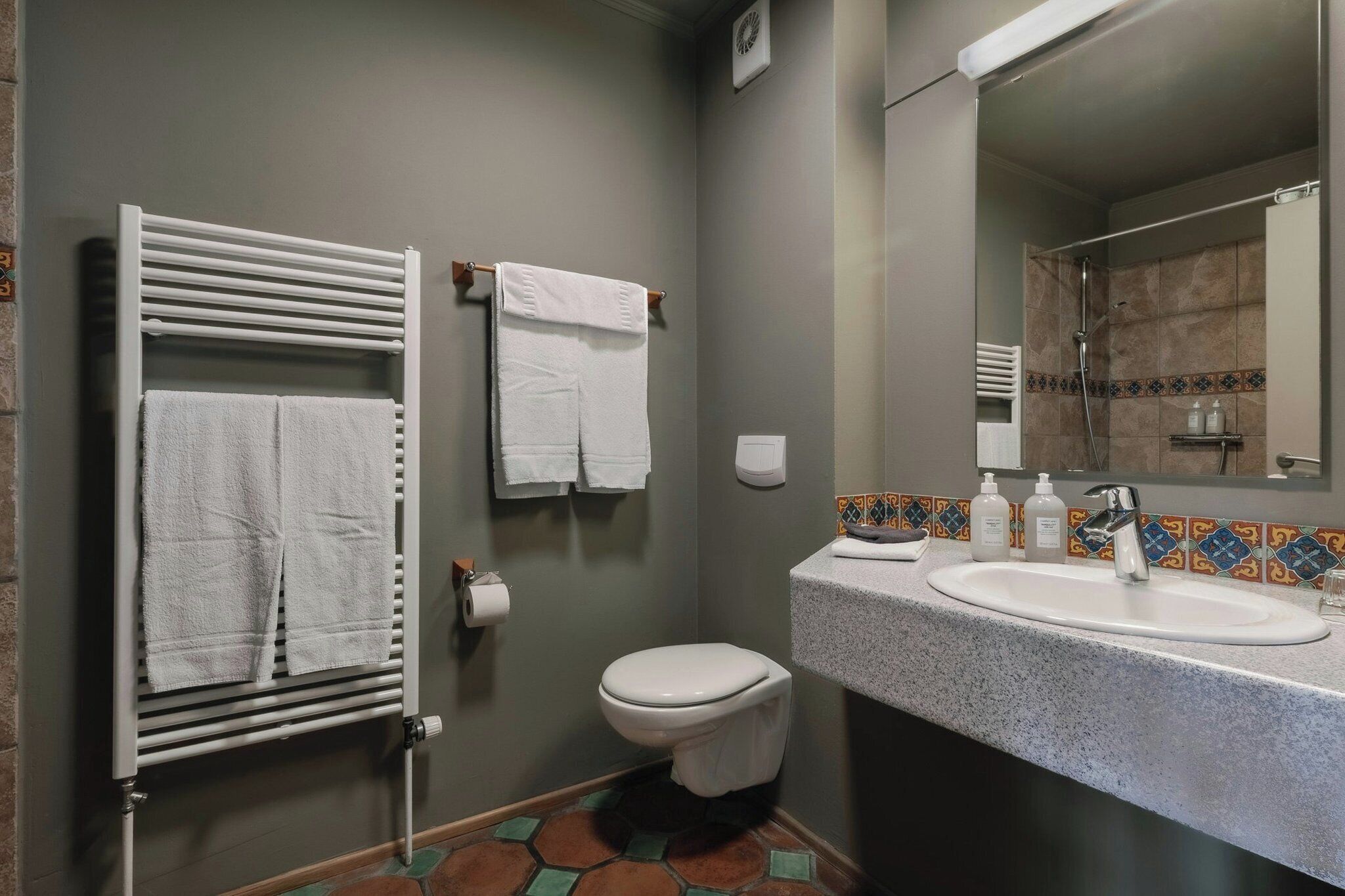 Un baño con inodoro blanco, un perchero con dos toallas, un lavamanos con espejo y una zona de ducha.