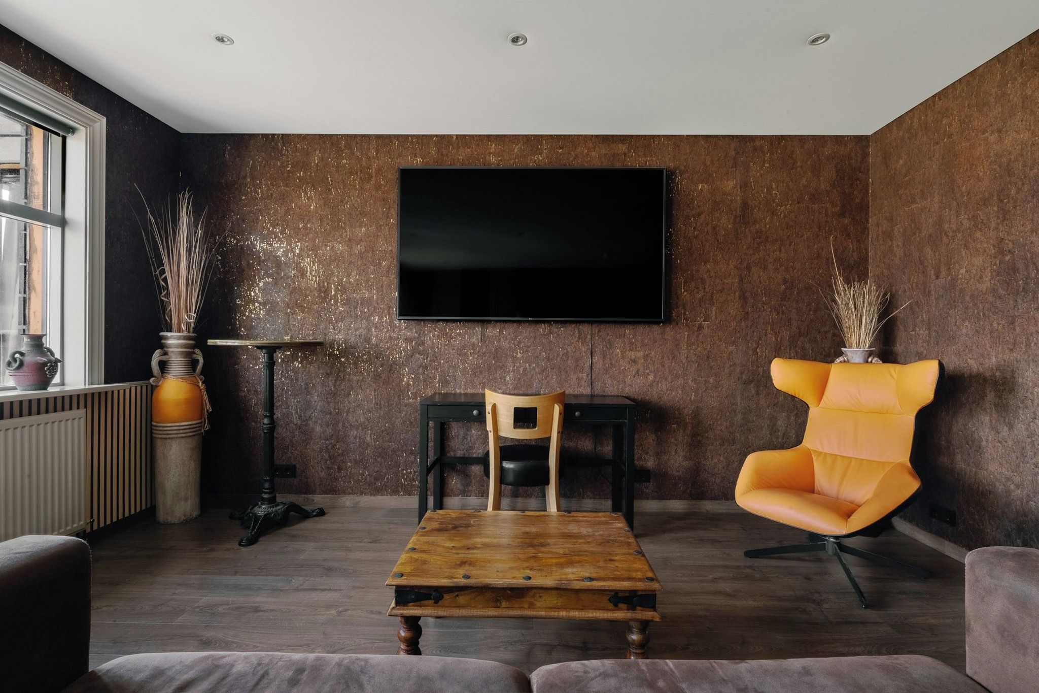 A modern living room with a large flat-screen TV mounted on the wall, a wooden coffee table, a yellow armchair, and a small wooden table with chairs.