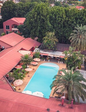 Una piscina rodeada de palmeras y sombrillas, con un edificio de techo rojo al fondo.