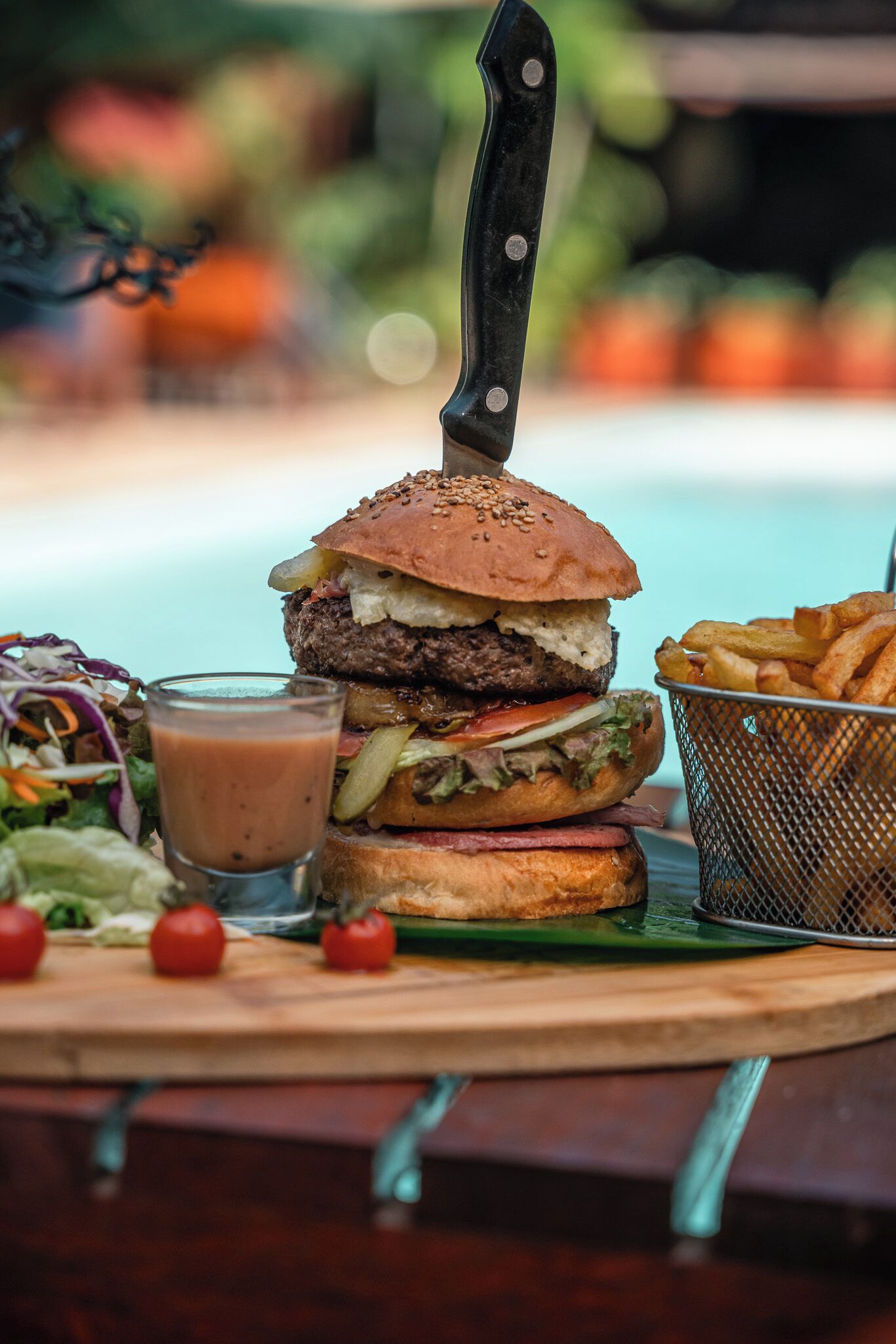 Hamburguesa con un cuchillo encima, un vaso de salsa, papas fritas y una ensalada sobre una tabla de madera.