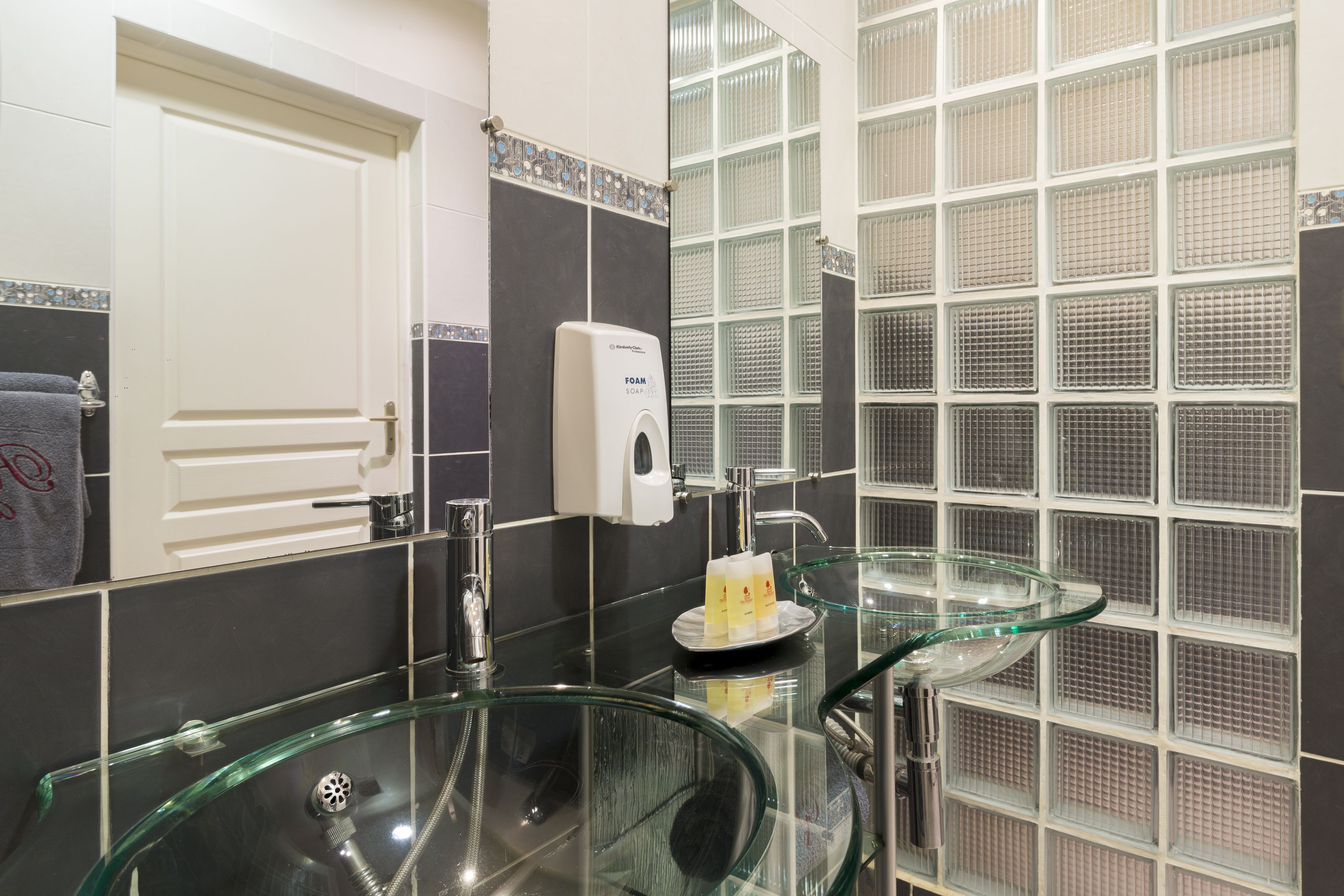 Een badkamer met een glazen wastafel, een zeepdispenser en een glazen schaal op het aanrecht.