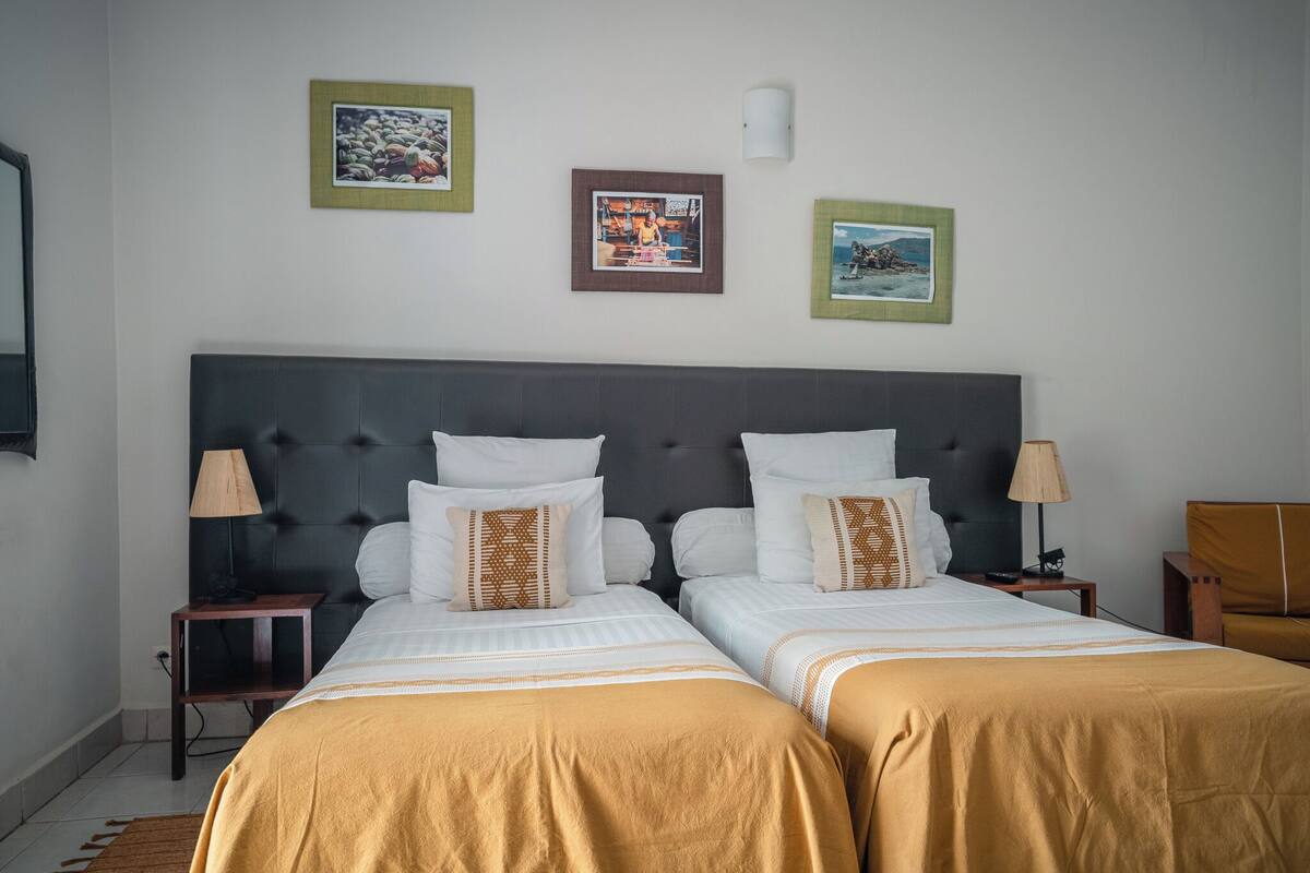 A hotel room with two beds, a large headboard, bedside tables with lamps, and framed pictures on the wall.