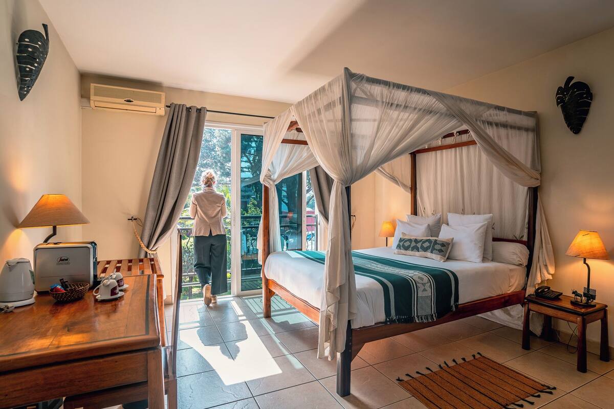 A bedroom with a four-poster bed, a desk with a lamp, and a view of the outdoors.
