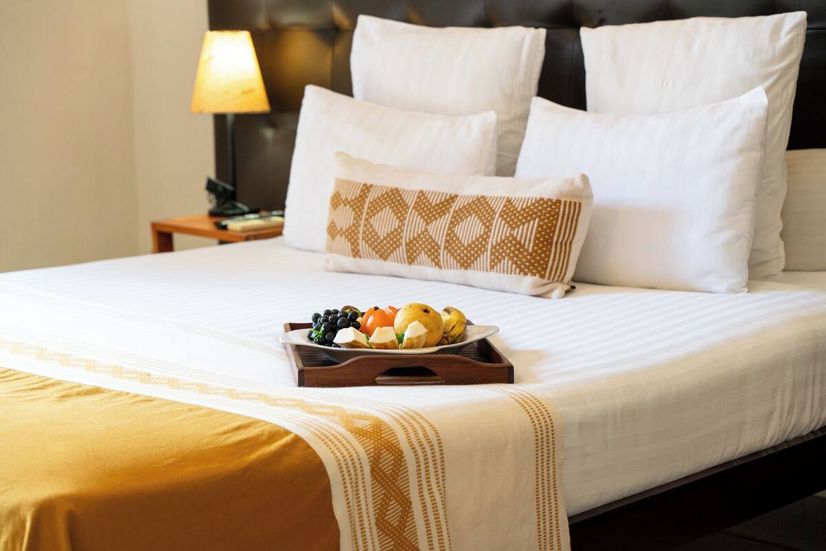 A neatly made bed with white linens, a tray of fruit, and a patterned pillow.