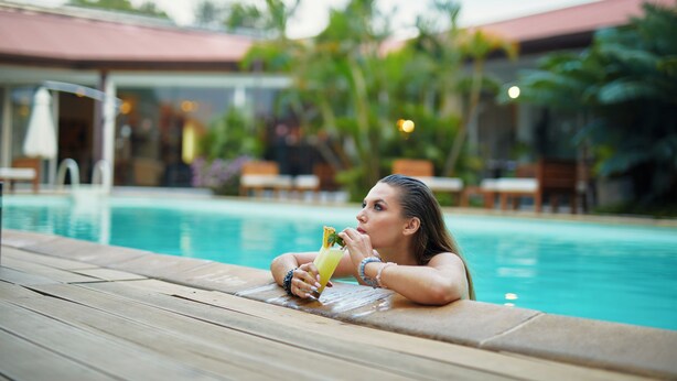 A woman relaxing by a pool with a drink, surrounded by greenery and a building with a red roof.