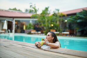 A woman relaxing by a pool with a drink, surrounded by greenery and a building with a red roof. - Relais des Plateaux (Antananarivo)