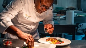A chef in a white uniform with "Bacon D" on the apron, preparing food in a kitchen with a hanging light fixture.