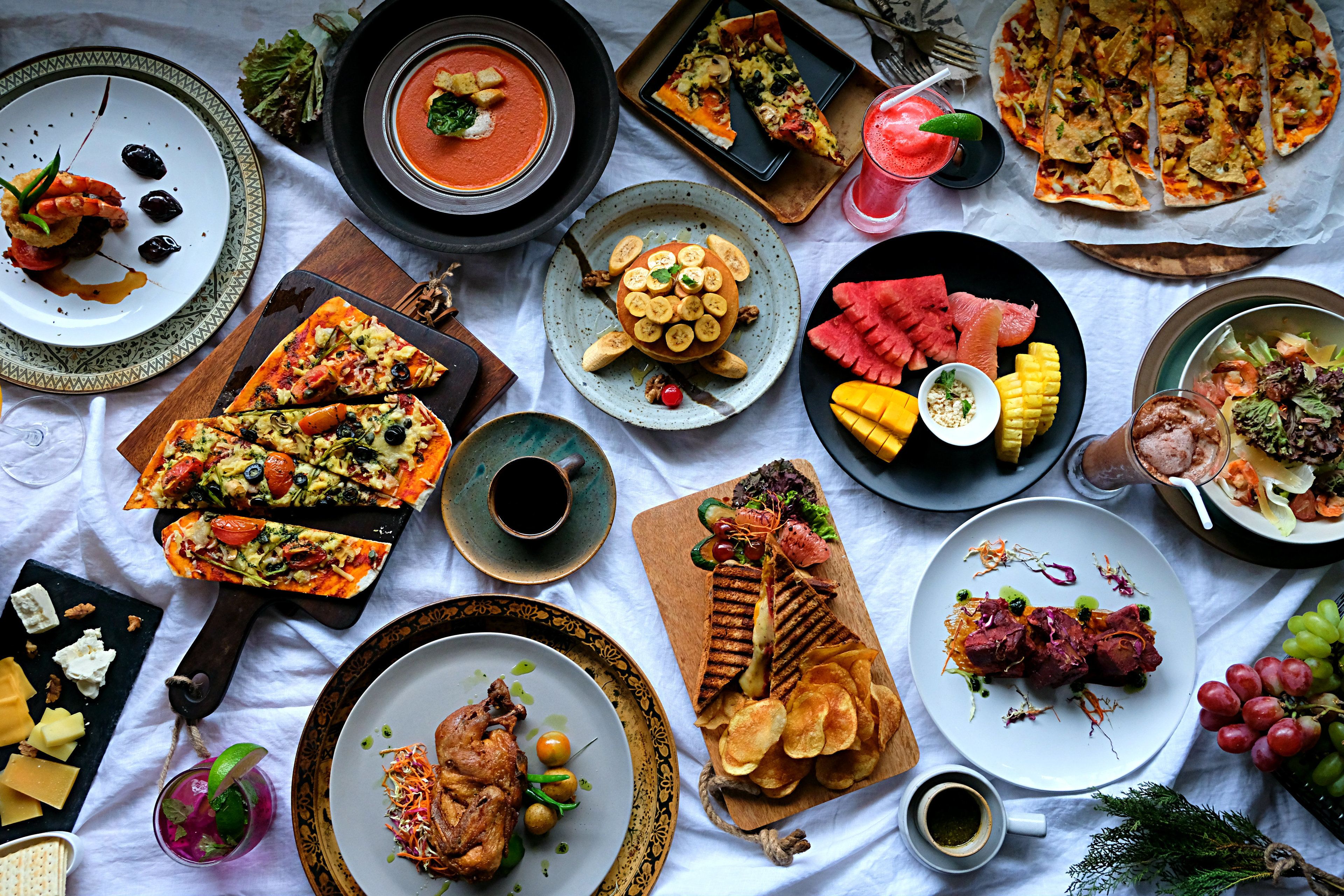 A variety of dishes including pizza, soup, and grilled meat, arranged on a table covered with a white cloth.