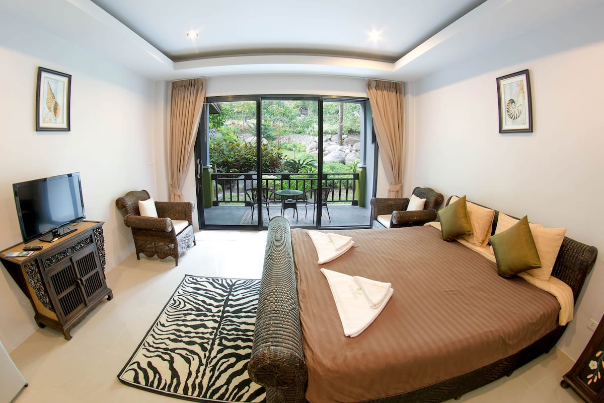 A spacious bedroom with a large bed, a television, a sofa, and a balcony with a view of greenery.