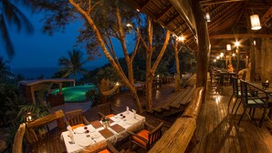 An outdoor dining area with wooden tables and chairs, surrounded by trees and overlooking a pool. - Tanote Villa (Koh Tao)