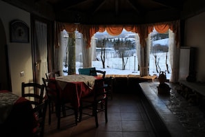 A dining area with a table set for four, chairs, a bench, and a view of a snowy landscape through large windows with curtains. - Rifugio del Pettirosso (Pescasseroli)
