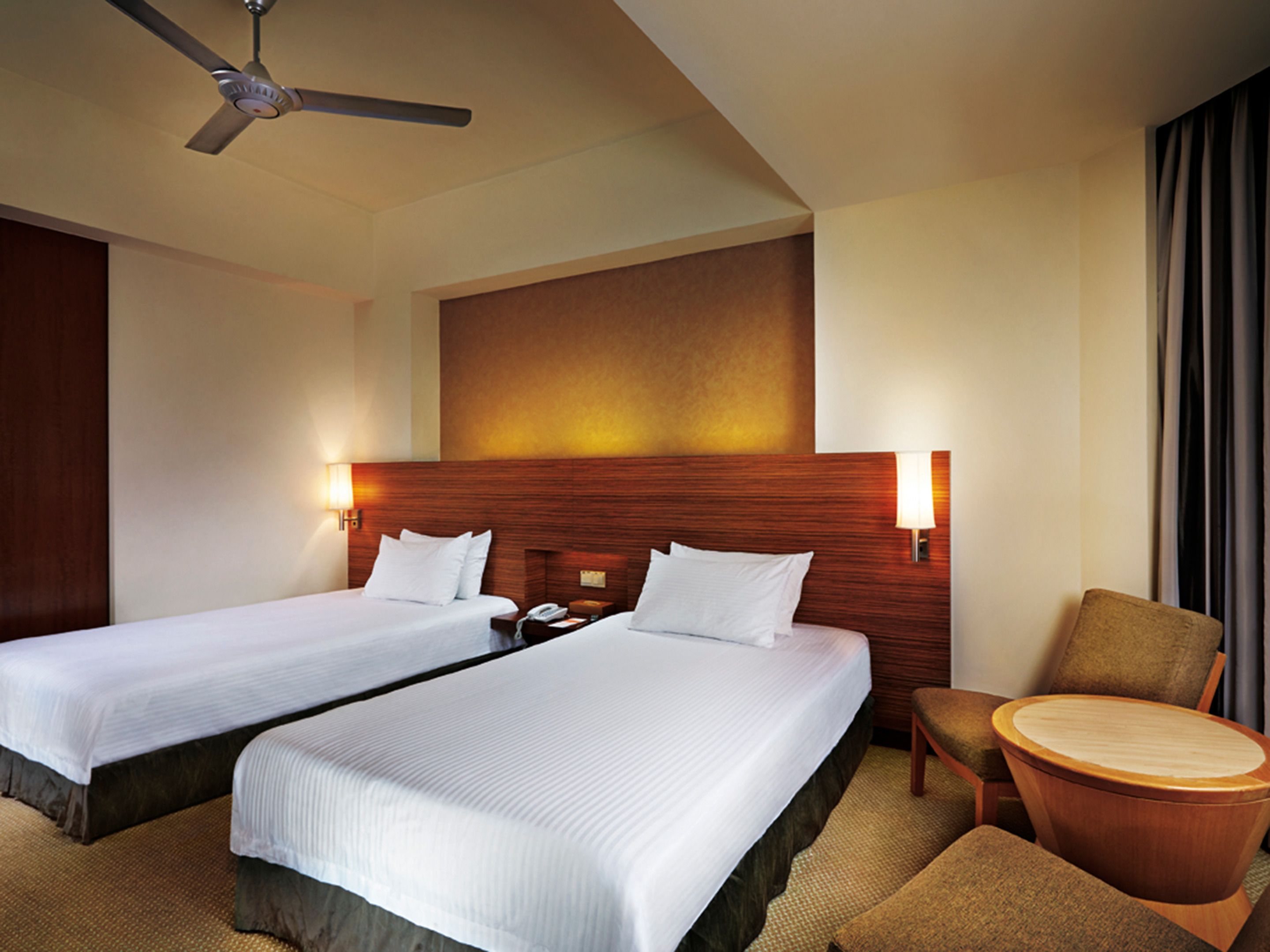 A hotel room with two beds, a ceiling fan, a wooden headboard, a nightstand, a chair, and a round table.