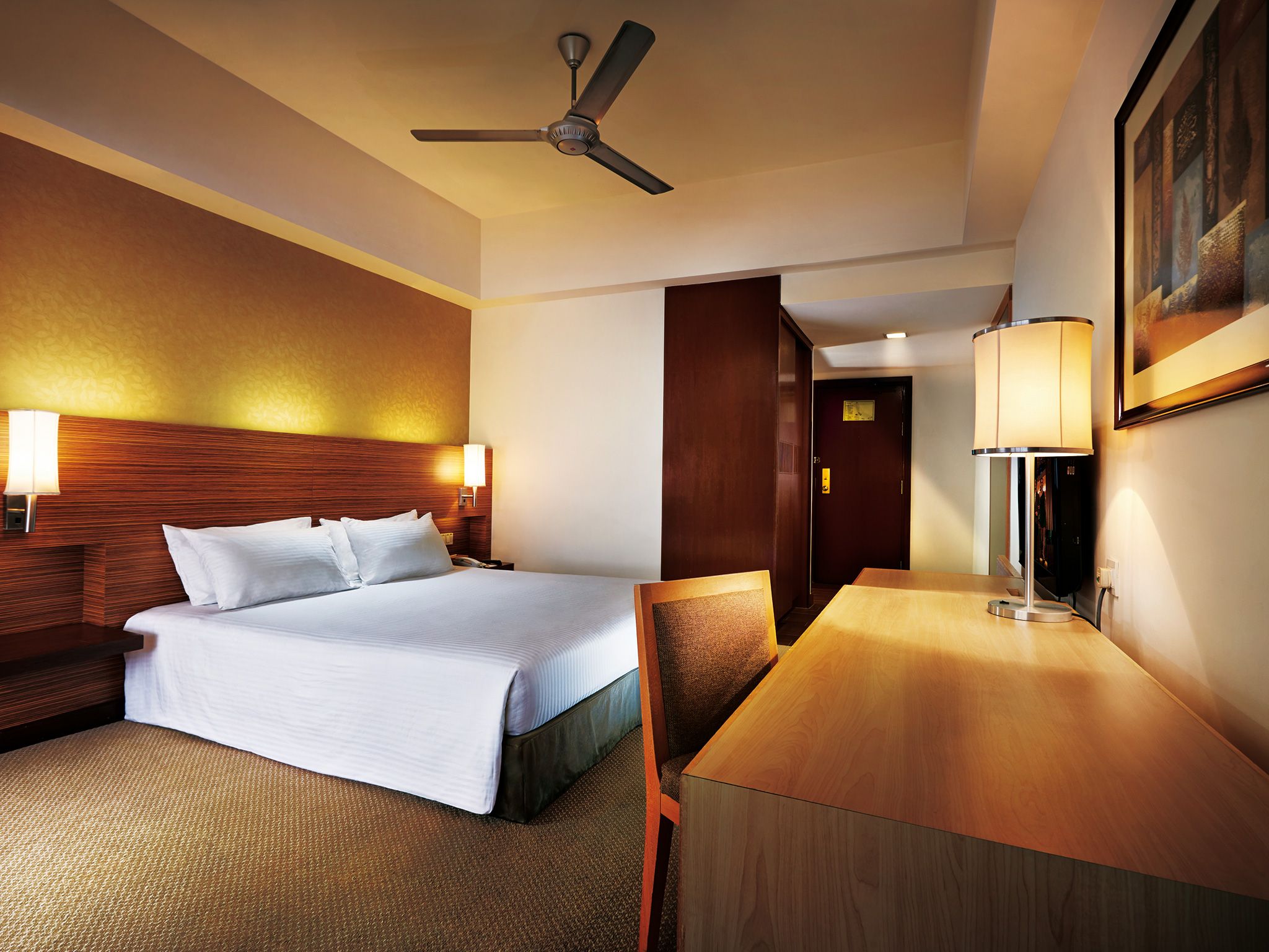 A hotel room with a bed, a desk, a chair, a ceiling fan, and two wall-mounted lamps.