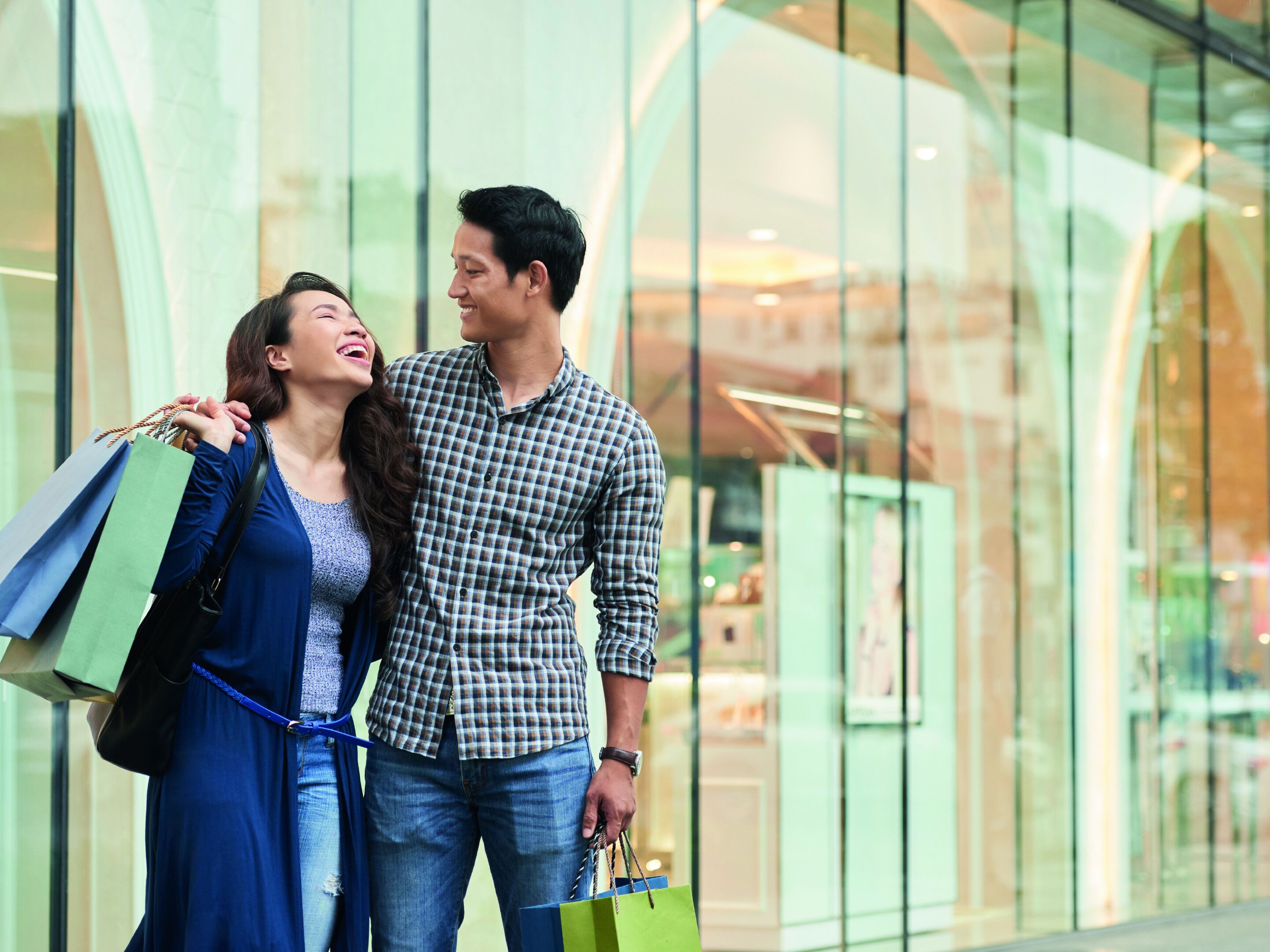 A couple holding shopping bags in front of a modern building with large glass windows.
