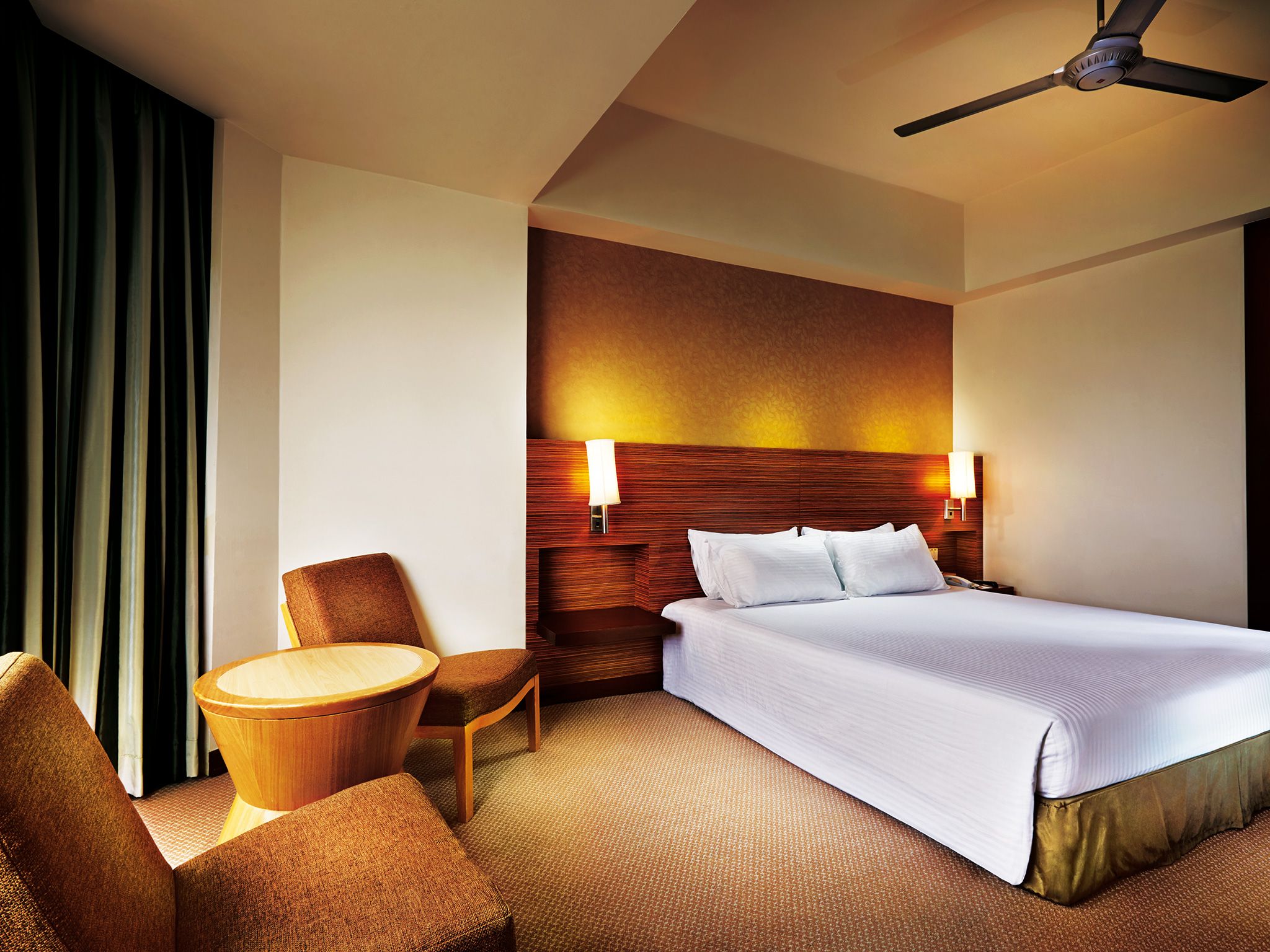 A hotel room with a bed, a wooden headboard, two wall-mounted lamps, a ceiling fan, a round wooden table, and a brown armchair.