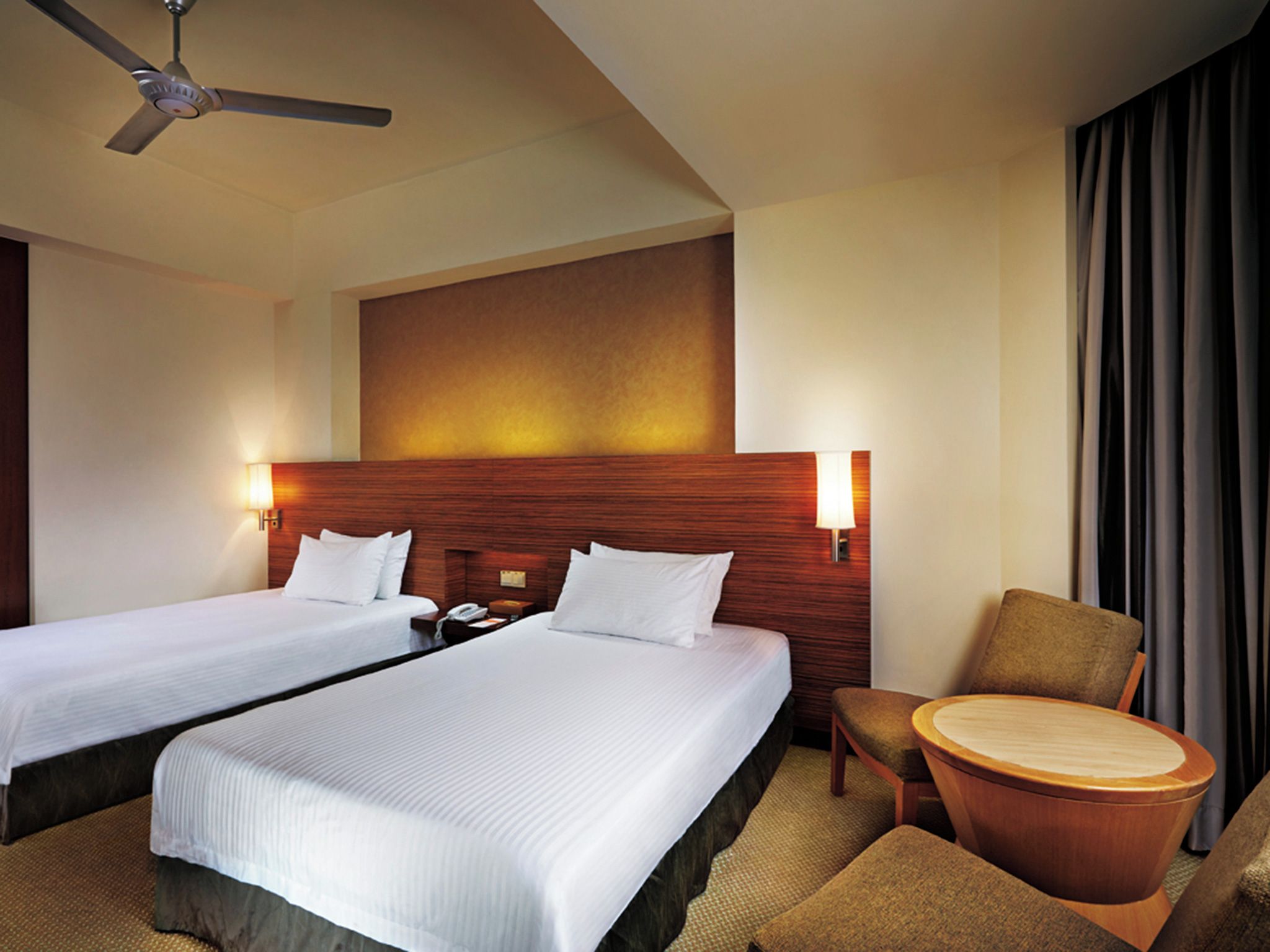 A hotel room with two beds, a wooden headboard, a chair, a round table, and a ceiling fan.